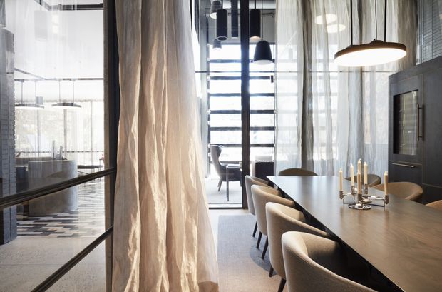 A refined contemporary dining area with a long dark timber dining table, upholstered dining chairs, sculptural pendant lighting, sheer floor-to-ceiling curtains, and expansive glazing. A sleek dark kitchen or living area is visible through the glazed partition on the left, while a tall window with horizontal framing anchors the background. The space has a sophisticated, softly illuminated atmosphere with strong vertical architectural elements.