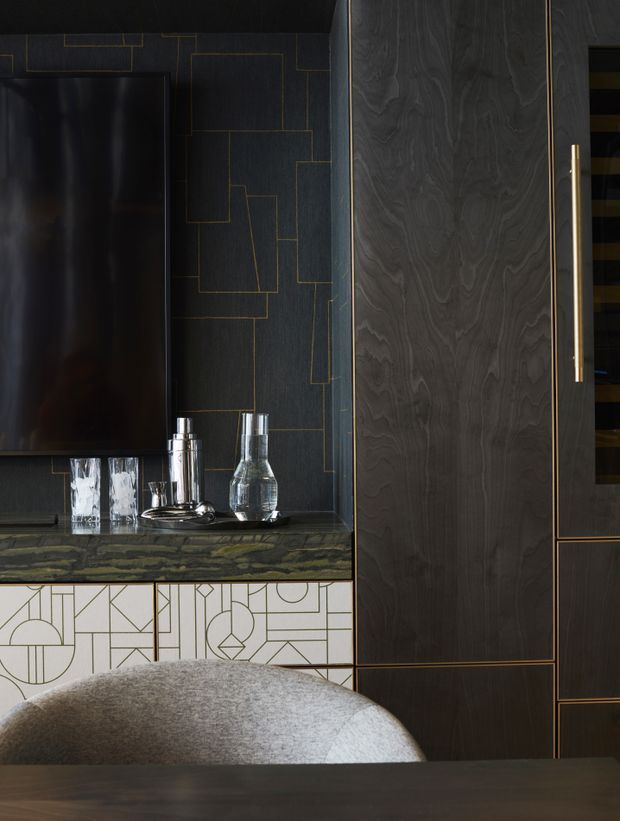 A moody, high-end entertainment or living-room joinery vignette featuring a wall-mounted black television, dark timber cabinetry with slim brass detailing, a dark stone or marble-look benchtop, and a geometric patterned cabinet front below. A curved upholstered chair and dark tabletop appear in the foreground, with glassware and decorative vessels arranged on the counter.