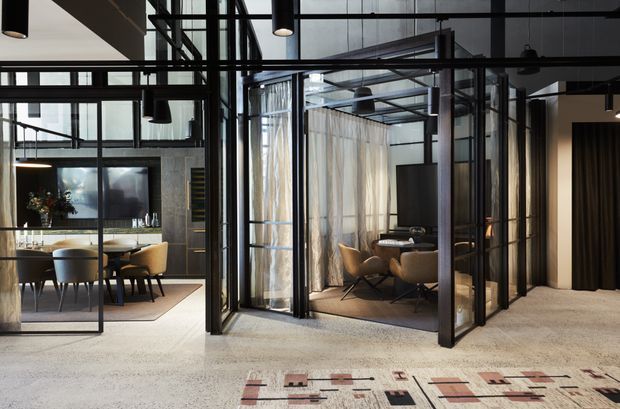 A sophisticated open-plan commercial or hospitality-style interior with black-framed glass partitions creating separate dining and meeting areas. The space features exposed structural elements, high ceilings, suspended black pendant lights, neutral furnishings, sheer curtains, polished concrete or terrazzo-like flooring, and a geometric area rug. Large internal glazing provides visual connection between the zones while maintaining separation.