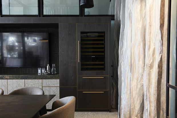A sophisticated dark-toned dining and entertainment area with a wall-mounted television, integrated dark timber cabinetry, a built-in wine fridge, patterned tiled backsplash, and a dark dining table with upholstered chairs. Sheer textured curtains cover tall glazing on the right, while black-framed upper windows and a black pendant light add an industrial-contemporary character.
