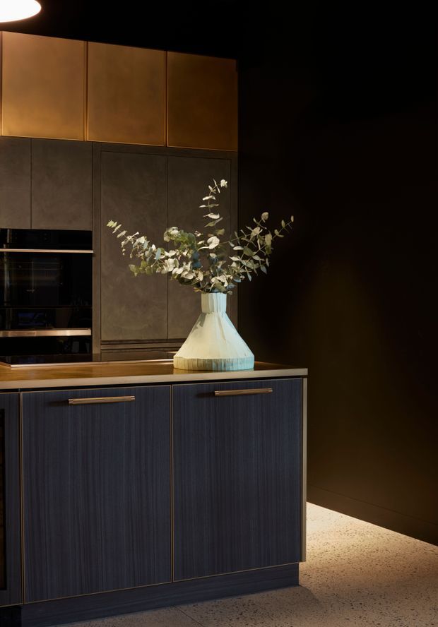A moody, close-up view of a contemporary kitchen featuring dark vertically textured lower cabinetry with brass-toned handles, bronze-brown upper cabinetry, integrated black appliances, and a warm wood-toned benchtop. A sculptural pale vase with eucalyptus branches sits on the counter, while dramatic low lighting highlights the cabinetry and speckled flooring.