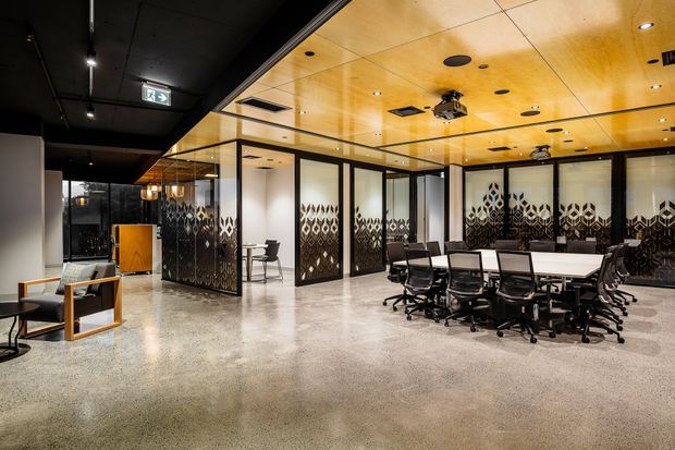A contemporary commercial office interior featuring a large open meeting or collaboration area, extensive black-framed glass partitions with patterned privacy film, a long white conference table surrounded by black office chairs, polished concrete flooring, and a warm timber-panel ceiling with integrated lighting and services. A darker lounge or reception area is visible at the left.