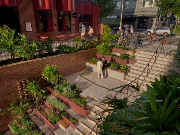 A landscaped urban courtyard and stepped public entrance beside a red brick commercial building. Broad tiled stairs and terraced raised garden beds lead between pedestrian areas, with lush planting, black metal railings, bicycles, and people using the space. The scene has a warm, contemporary streetscape character.
