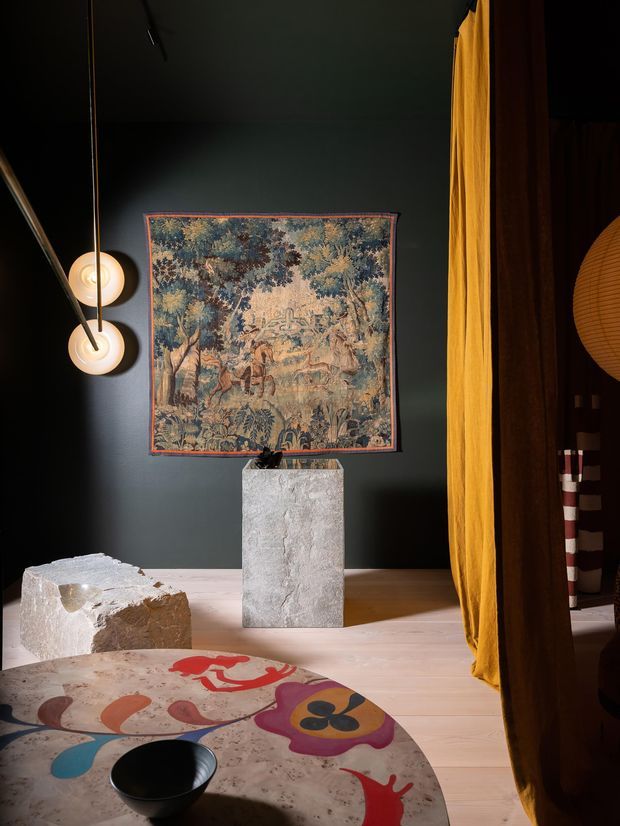 A moody, art-filled interior corner with deep charcoal walls, warm timber flooring, a vintage tapestry, sculptural stone planters, globe pendant lights, and a mustard-gold curtain. A colourful rounded table with abstract painted motifs and a black bowl occupies the foreground, creating an eclectic, gallery-like atmosphere.