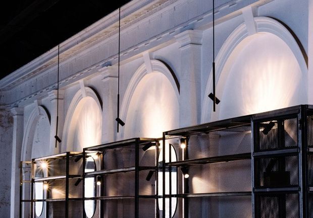 Interior architectural view of a grand, heritage-style space with a long white plaster wall articulated by repeated tall arched recesses and elaborate classical cornice detailing. Contemporary black metal mesh shelving or display structures occupy the foreground, with slim suspended black pendant lights and warm concealed illumination washing the arches. The composition contrasts ornate historic fabric with industrial-modern insertions.