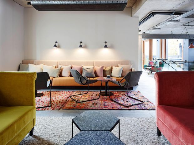 A stylish open-plan lounge with a long peach-toned sofa, sculptural dark lounge chairs, colourful patterned rug layered over a light carpet, and mustard and red armchairs in the foreground. The room has exposed concrete ceilings, visible services, black wall sconces, and glazing opening toward a bright adjoining workspace or dining area.