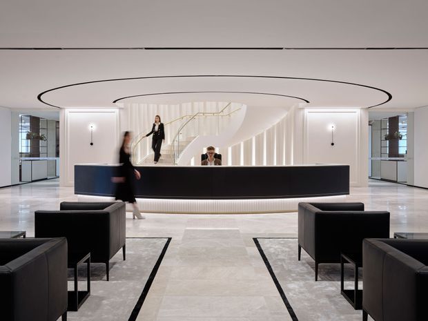 A refined contemporary hotel or commercial reception lobby viewed head-on. A long, dark reception desk sits beneath a large oval recessed ceiling feature with black edging. Behind it, a sculptural curved staircase and vertical illuminated slats create a dramatic architectural backdrop. The spacious lobby has pale stone-look flooring, white walls, mirrored or glazed side panels, and paired black lounge chairs arranged symmetrically in the foreground.