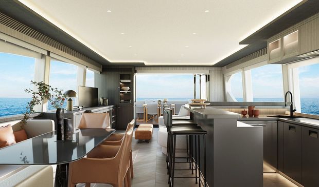 A refined open-plan coastal interior combining a kitchen, dining area, and living space. Floor-to-ceiling glazing surrounds the room and offers expansive ocean views. The kitchen features dark built-in cabinetry, a large pale island with bar seating, integrated lighting, and a stainless-steel tap. A round dark dining table with upholstered chairs sits beside a compact living area, while warm timber and neutral finishes create a polished contemporary feel.