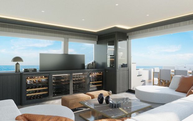 Luxurious contemporary living room with floor-to-ceiling ocean-facing glazing, a large sectional sofa, glass coffee table, and dark built-in cabinetry incorporating wine refrigerators and display storage. The room opens to a bright outdoor terrace with dining furniture and expansive sea views.