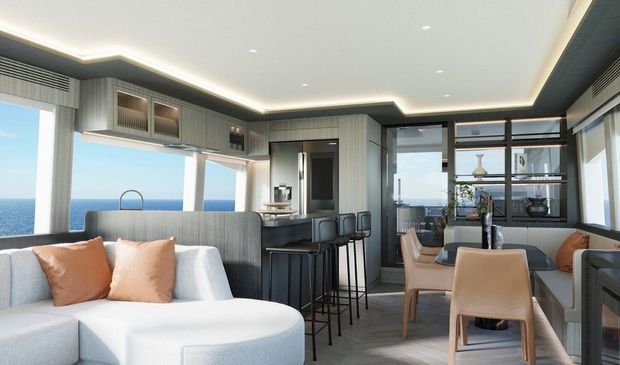 A bright, sophisticated open-plan coastal interior combining a living room, kitchen, and dining area. The space features floor-to-ceiling glazing with ocean views, a curved light upholstered sofa, dark textured cabinetry, a compact island with bar stools, a dining table with tan upholstered chairs, integrated shelving, and concealed LED ceiling lighting.
