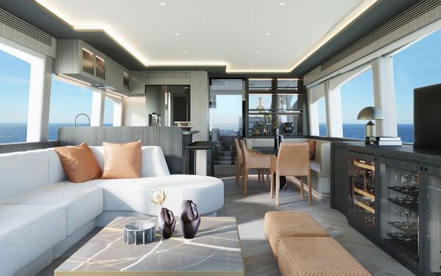 A bright, contemporary open-plan living and dining space with a compact kitchen, extensive floor-to-ceiling ocean-facing glazing, and a refined grey-toned interior. The room features a large pale sectional sofa, textured ottoman seating, a stone-look coffee table, timber dining chairs, dark grey built-in cabinetry, integrated lighting, and a drinks cabinet. Ocean views are visible through the windows.