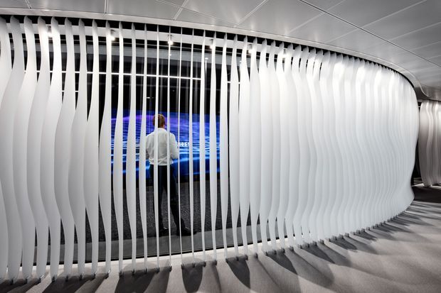 A futuristic commercial interior featuring a curved partition made from repeated white vertical wavy fins suspended from a metallic ceiling track. The semi-transparent screen encloses or separates a display area with blue illuminated graphics, while dramatic directional lighting casts elongated shadows across the grey floor. The space has a sleek, high-tech and immersive character.