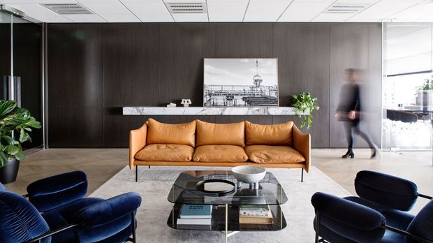 A refined commercial reception or lounge area with a caramel leather three-seat sofa, deep navy upholstered lounge chairs, and a low glass coffee table. Dark timber wall panelling forms the backdrop, with a slim stone or light-coloured ledge, framed artwork, potted plants, and a bright glazed office area to the right. The ceiling uses a suspended modular grid with recessed linear lighting, while the floor has a light neutral hard finish. The composition has a polished, contemporary corporate interior character.