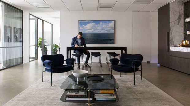A refined contemporary office lounge and meeting area with dark blue lounge chairs arranged around a low glass coffee table, a high communal table with stools, pale timber flooring, a large neutral rug, and floor-to-ceiling glazed partitions. Fixed dark timber joinery and a marble-look feature wall with open shelving are visible on the right. The space has a suspended acoustic ceiling and abundant natural light.