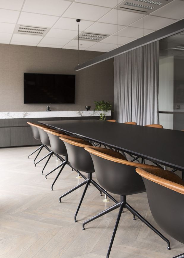 A contemporary boardroom or meeting room with a long matte-black conference table surrounded by modern chairs with dark upholstered shells, black star-shaped metal bases, and curved timber armrests. The room features light herringbone timber flooring, dark grey built-in cabinetry, a white marble-look benchtop or credenza surface, a wall-mounted television, grey curtains, and a suspended linear light fitting. A suspended acoustic tile ceiling with recessed fluorescent-style panels is visible overhead.
