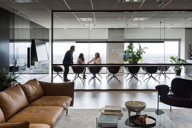 A bright contemporary office lounge and meeting area with floor-to-ceiling glazed partitions and expansive city views. A long conference table with black chairs sits beside the windows, while a tan leather sectional, black lounge chair, glass coffee table, indoor plants, and a wall-mounted screen furnish the foreground. The office has a suspended acoustic ceiling and polished, minimal finishes.