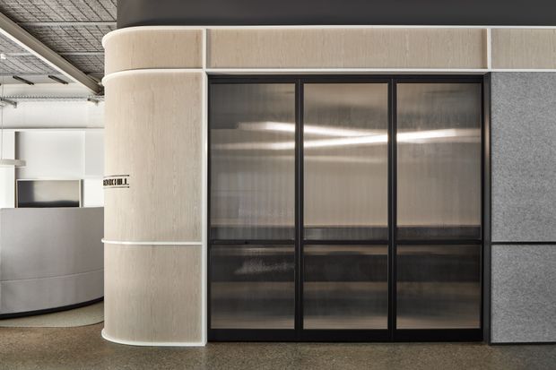 Contemporary commercial interior featuring a curved light wood-toned reception or partition wall, black-framed translucent glass doors, grey acoustic wall panels, and polished concrete or terrazzo flooring. The space has an industrial ceiling grid and refined minimalist detailing.