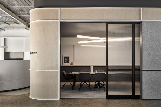 Contemporary commercial interior showing a glazed meeting room enclosed by black-framed partitions, with a central conference table and dark chairs. The room is integrated into a softly curved, light-toned built-in wall and reception-like enclosure, alongside grey acoustic partition panels. Exposed services and a grid ceiling are visible in the surrounding office environment, with linear pendant lighting inside the meeting room.