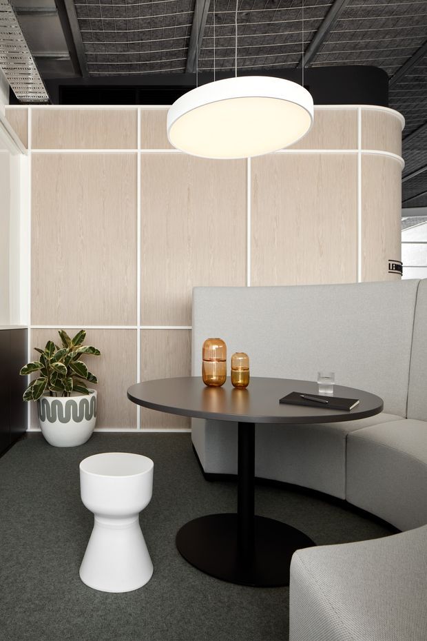 A contemporary commercial lounge or breakout area with a curved upholstered booth, round black pedestal table, white sculptural side stool, potted plant, amber glass vessels, and a large suspended circular pendant. Pale timber-look wall panels with white grid framing form a curved partition, while a dark exposed services ceiling and charcoal carpet create contrast.