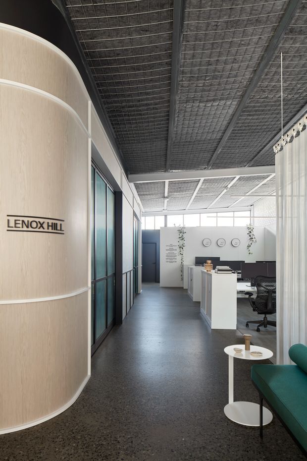 A bright contemporary office reception and workspace interior with a curved pale timber feature wall bearing the “LENOX HILL” sign. The space has exposed dark metal ceiling grids and services, polished dark flooring, glazed meeting-room partitions, white workstations, black office chairs, sheer curtains, and a teal upholstered lounge seat with a small round side table. Natural light enters through high-level glazing, creating a refined industrial-commercial atmosphere.