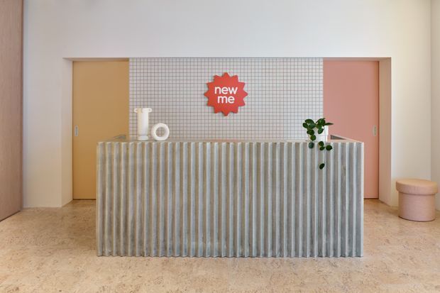 A playful contemporary reception or service counter is centered in the frame. The foreground features a large fluted vertical-panel counter in pale grey, set against a small square-grid tiled wall with a red “new me” sign. Warm mustard and blush-pink doors flank the tiled feature wall, while a small plant, sculptural white objects, and a rounded upholstered stool add decorative accents. The space has a warm, softly lit, highly graphic interior fit-out.
