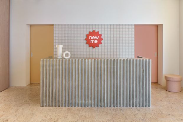 A playful contemporary reception area featuring a large fluted ribbed counter in the foreground, a small square-grid tiled feature wall behind it, and pastel mustard and pink door panels. The pale terrazzo-like floor, warm off-white walls, and minimal decorative objects create a bright, graphic interior.