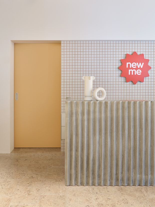A stylised commercial reception or service counter with a warm mustard sliding door, pale pink grid-pattern tiled wall, and a tall fluted grey-beige counter front. Small sculptural objects sit on the counter, alongside a bright red graphic sign reading “new me.” The space has a playful contemporary fit-out with a terrazzo-like beige floor and soft neutral lighting.