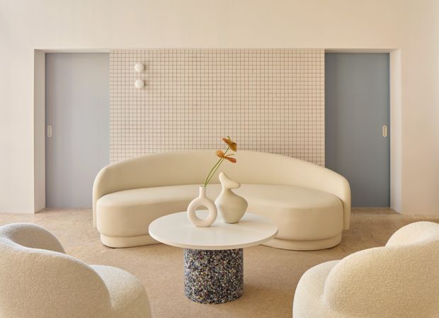 A softly lit, minimalist lounge or reception space featuring a curved cream sofa and rounded boucle lounge chairs arranged around a low circular coffee table. The table has a speckled cylindrical pedestal, while sculptural ceramic vases and a single dried stem provide understated decoration. Behind the seating is a central grid-pattern tiled wall flanked by two recessed grey doors or panels. The palette is warm and neutral with rounded, tactile forms and a refined contemporary feel.