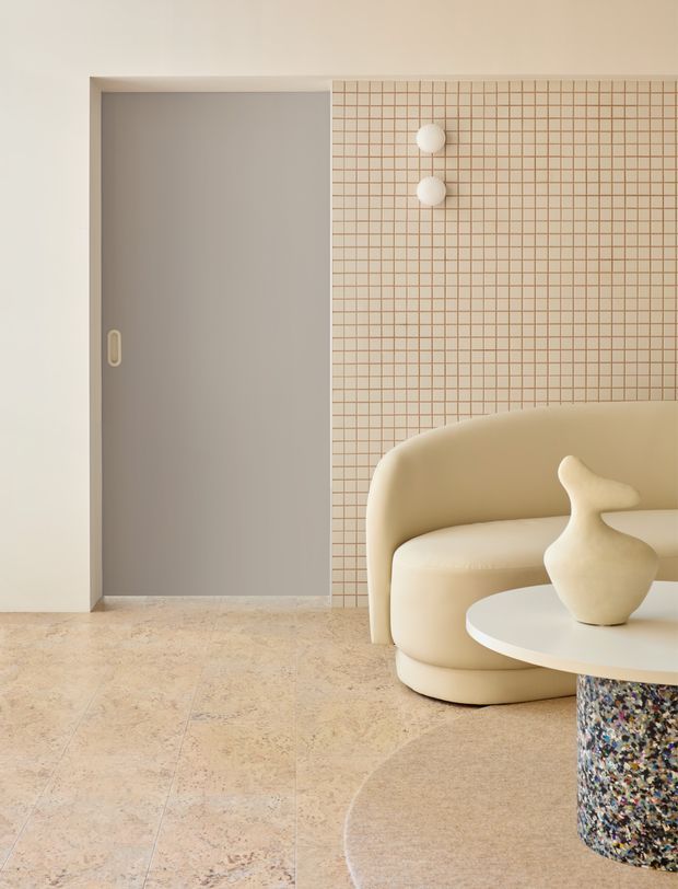 A softly lit contemporary living room with a sculptural cream curved sofa, round white coffee table, abstract ceramic vase, and a terrazzo-like table pedestal. The room features a pale beige tiled floor, a warm neutral wall, a recessed grey sliding door, and a grid-patterned tiled feature wall with two spherical wall lights. The overall composition is refined, minimalist, and softly organic.