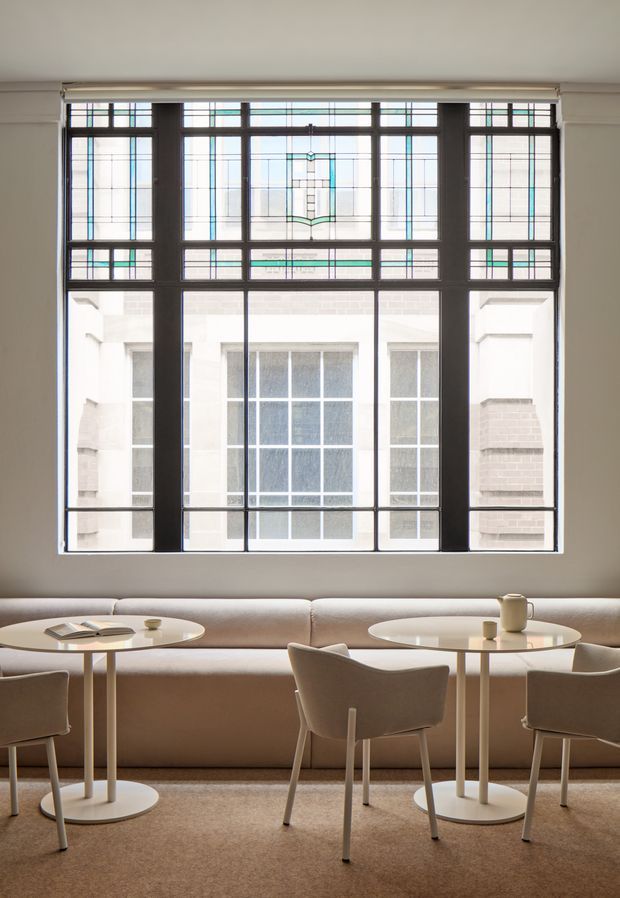 A refined, softly lit lounge or hospitality seating area with a long upholstered banquette beneath a large decorative leaded-glass window. Two small round pedestal tables are paired with upholstered dining-style chairs, creating an elegant café or hotel lobby setting. The palette is warm and neutral, with beige carpet, taupe upholstery, cream walls, dark window mullions, and subtle stained-glass detailing.