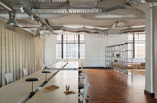 A bright open-plan commercial interior with long communal tables, chairs, black table lamps, exposed ductwork and cable trays, tall industrial-style windows, a full-height ribbed curtain, open metal shelving, and warm herringbone timber flooring. The space has a refined warehouse-office character with a neutral palette and abundant natural light.