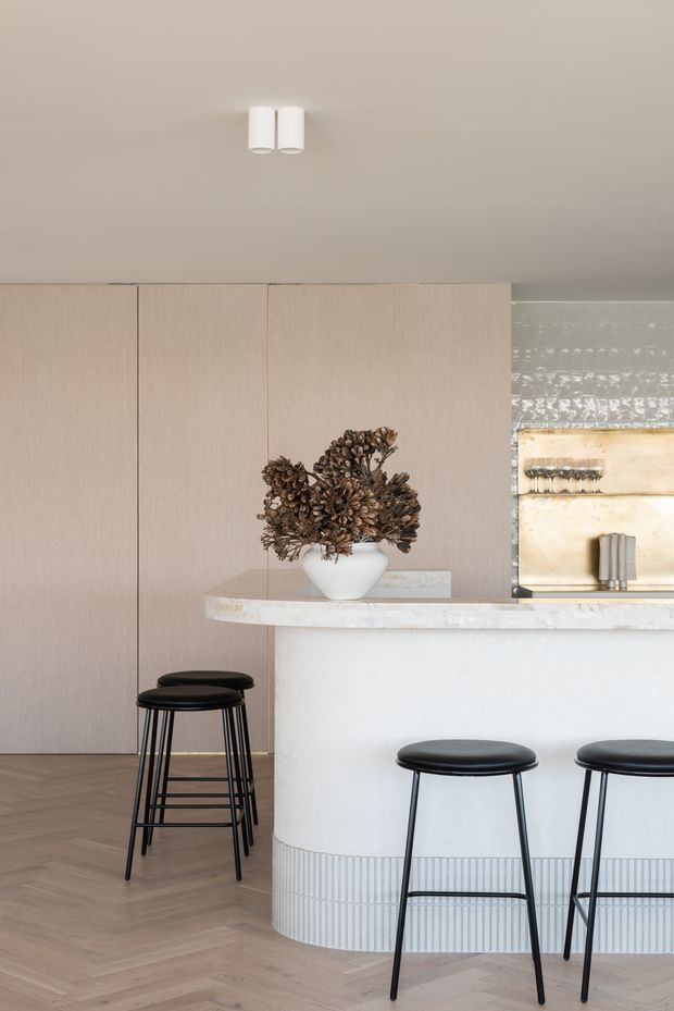 A refined contemporary kitchen and breakfast bar with a curved white island, pale stone countertop, fluted tile base, light timber wall panelling, and herringbone timber flooring. Black bar stools provide contrast, while a sculptural floral arrangement sits on the counter. A textured tiled backsplash and framed artwork are visible behind the island.