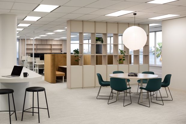 A bright, contemporary open-plan commercial office interior with a co-working or breakout area. The space features a white curved reception or kitchenette counter with black stools, a round meeting table surrounded by dark teal chairs, pale timber open shelving and partition joinery, indoor plants, expansive windows, and a suspended spherical pendant light. A suspended grid ceiling with recessed fluorescent panels and light carpet create a clean, functional workplace atmosphere.