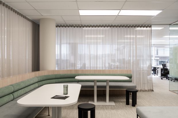 Contemporary commercial breakout lounge with a curved built-in sage-green banquette, pale wood wall-height backing, white rounded tables, and black stools. Sheer vertical blinds, a suspended grid ceiling with fluorescent panel lighting, carpeted flooring, and glazed office spaces create a bright, open-plan workplace setting.