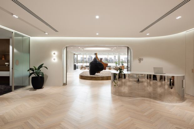 A refined contemporary reception or entry lobby with pale herringbone timber flooring, curved cream walls, recessed linear ceiling vents and downlights, and a sculptural curved stone reception desk. A glazed opening leads to a bright lounge or gallery area with city views, while built-in shelving and decorative lighting add detail to the lobby.