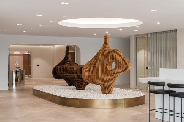 A refined contemporary interior featuring a large sculptural wooden fish installation on a raised oval plinth filled with white stones. The space has pale herringbone timber flooring, softly curved architectural openings, a warm neutral palette, recessed ceiling lighting, and a white kitchen or bar counter with black stools in the foreground. A glass-partitioned room and reflective surfaces extend the view into the background.