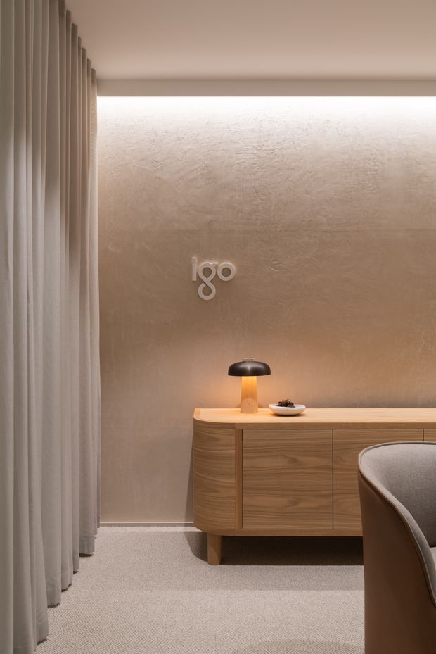 A refined contemporary interior vignette featuring a rounded timber sideboard with integrated drawers, a textured taupe plaster wall, soft floor-to-ceiling curtains, and warm indirect lighting. A small table lamp and ceramic bowl sit on the sideboard, while part of a curved upholstered chair is visible in the foreground. The atmosphere is calm, warm, and understated.