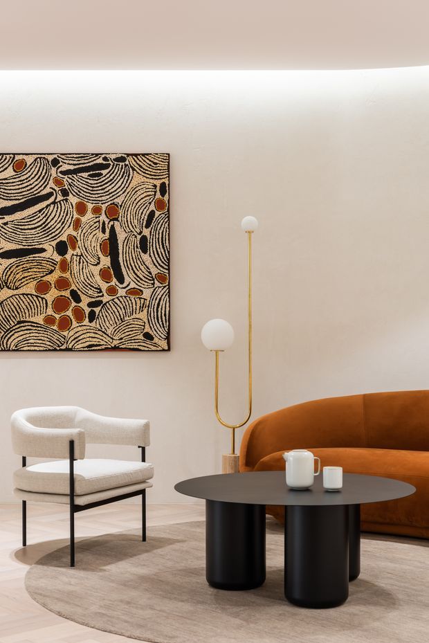 A warmly styled living room with a sculptural rust-orange curved sofa, cream upholstered lounge chair, black cylindrical coffee table, and brass-and-globe floor lamp. The room has softly textured off-white walls, pale timber flooring, a large neutral round rug, and a bold abstract artwork with black, cream, and ochre organic patterns.