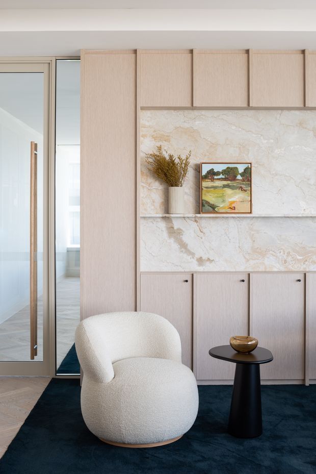 A refined residential interior vignette featuring a pale timber built-in cabinetry wall with upper and lower storage, a large veined stone feature surface and display shelf, and a glazed timber-framed door or partition at left. A sculptural off-white boucle lounge chair and small black pedestal side table sit on a deep navy rug, creating a calm, contemporary seating area. A framed landscape artwork and dried botanical arrangement add restrained decorative detail.