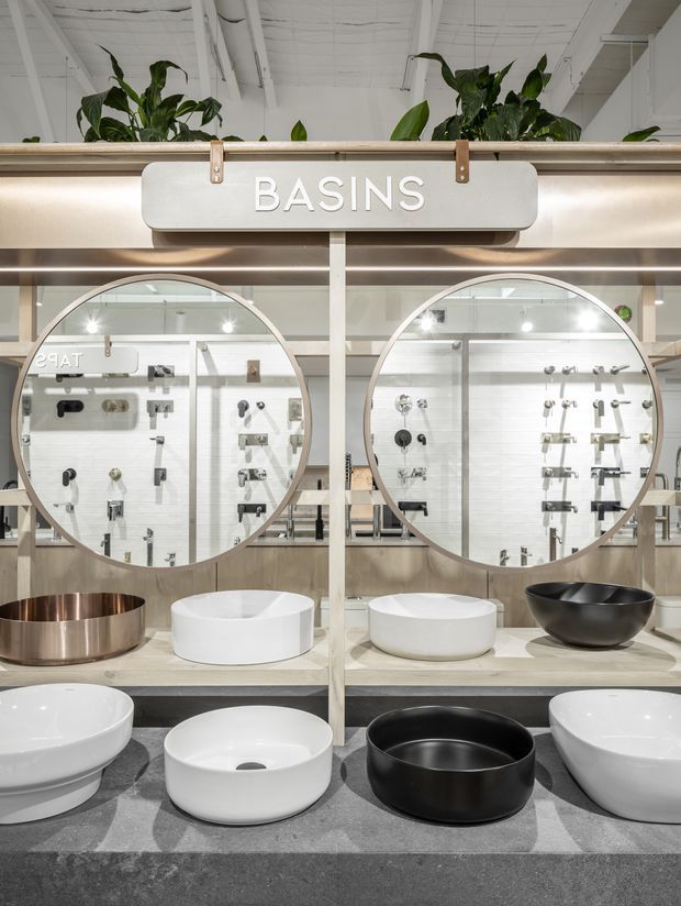 A bright bathroom showroom display focused on countertop basins. Multiple round and oval ceramic basins in white and black, along with a metallic copper-toned basin, are arranged on stone-look display counters. Large circular mirrors reflect rows of bathroom tapware and fixtures, while a prominent overhead sign reads “BASINS.” The space has light neutral finishes, integrated lighting, exposed white ceiling structure, and indoor plants.