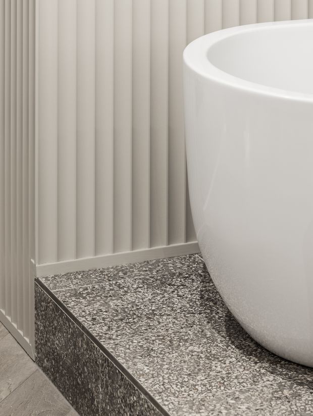 Close-up detail of a contemporary bathroom featuring a white freestanding bathtub set on a raised terrazzo platform. The surrounding walls have vertical fluted panelling in a soft neutral tone, with grey-and-white terrazzo flooring and step surfaces.