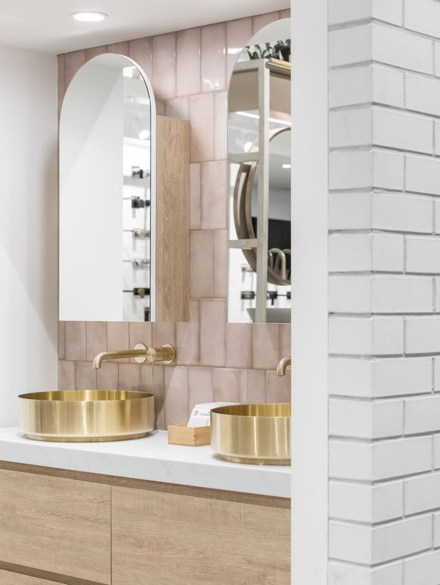 A bright contemporary bathroom vanity with twin round brushed-gold vessel basins, wall-mounted gold faucets, pale timber cabinetry, white countertop, and glossy blush-pink rectangular wall tiles. Arched mirrors and mirrored cabinetry add depth, while a white-painted brick partition frames the right side.