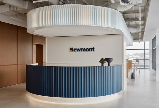 A contemporary commercial reception area featuring a large curved reception desk with deep blue vertically ribbed cladding, a white curved feature canopy above, and a pale stone or concrete floor. The Newmont logo is mounted on a light wall behind the desk. Exposed white mechanical services and ductwork create an open ceiling, while timber wall panelling and extensive glazing are visible in the surrounding office.