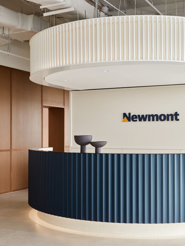 A contemporary commercial reception area featuring a large curved reception desk clad in vertically ribbed dark blue panels, a cream tiled plinth, and a pale solid-surface counter. Behind it is a curved cream wall with the Newmont logo, framed by warm wood wall panelling. An elliptical mezzanine or balcony with evenly spaced white vertical balusters projects overhead, while exposed services and suspended linear lighting remain visible in the high ceiling.