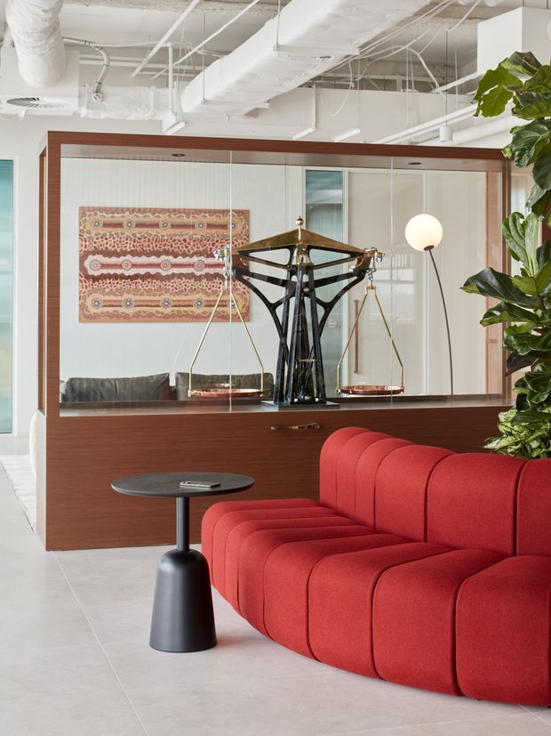 A sophisticated contemporary lounge or reception area featuring a vivid red modular sofa, a small black pedestal side table, and a large dark timber-framed mirrored built-in display or storage wall. A sculptural balance scale sits in front of the mirror, while a large leafy indoor plant adds greenery. The space has a bright open-plan commercial feel with exposed white services and ducts across the ceiling, pale flooring, and an Aboriginal-inspired patterned artwork reflected or mounted behind the display.