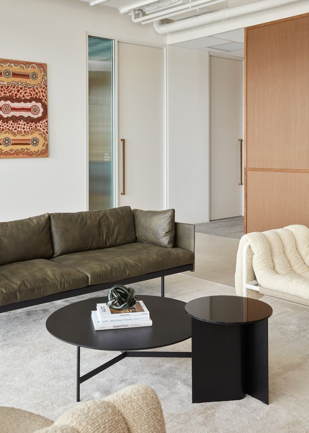 Contemporary office lounge with a dark leather sofa, sculptural black coffee tables, boucle lounge chairs, and a pale rug. The space has white walls, exposed white services and ceiling panels, glazed and solid interior doors with timber handles, and a warm timber feature wall. Aboriginal artwork adds colour to the otherwise muted, refined palette.