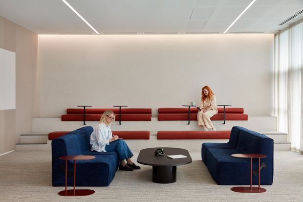 A bright contemporary commercial lounge or waiting area with tiered red upholstered bench seating against a pale wall, deep blue armchairs and sofas, and low dark coffee tables. Two people are seated in the space. Floor-to-ceiling sheer curtains line the right side, while linear recessed ceiling lighting creates a clean, refined atmosphere.