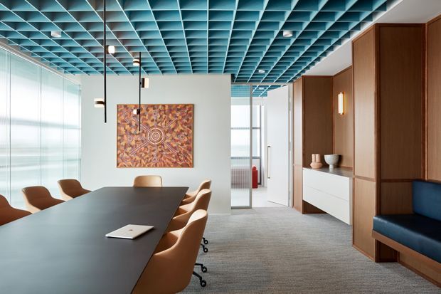 Contemporary commercial meeting room with a long dark conference table surrounded by tan upholstered chairs, extensive floor-to-ceiling glazing, and a striking turquoise geometric coffered ceiling grid. A timber-paneled built-in storage wall with white drawers and integrated blue upholstered bench seating lines the right side. The space has carpet flooring, minimalist pendant and wall lighting, and colourful artwork on a pale wall.