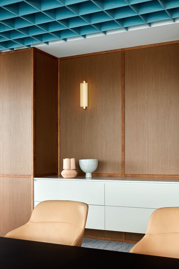 A refined mid-century-inspired interior with a dark wood-paneled feature wall, integrated white storage cabinetry, and two tan upholstered lounge chairs around a dark table. A sculptural wall sconce and small ceramic objects sit against the panelled backdrop, while a turquoise coffered or geometric open-grid ceiling creates a strong contrasting feature. The space appears to be a sophisticated office, meeting room, or lounge.