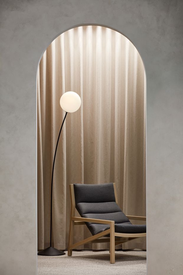 A refined interior vignette framed by a tall arched opening. Textured grey plaster walls surround a full-height opening lined with warm beige pleated curtains. A sculptural black floor lamp with a glowing spherical shade stands beside a dark charcoal upholstered lounge chair with a light timber frame and matching footrest, set on a pale textured carpet. The composition is quiet, warm, and minimalist with strong vertical lines and a curved architectural detail.